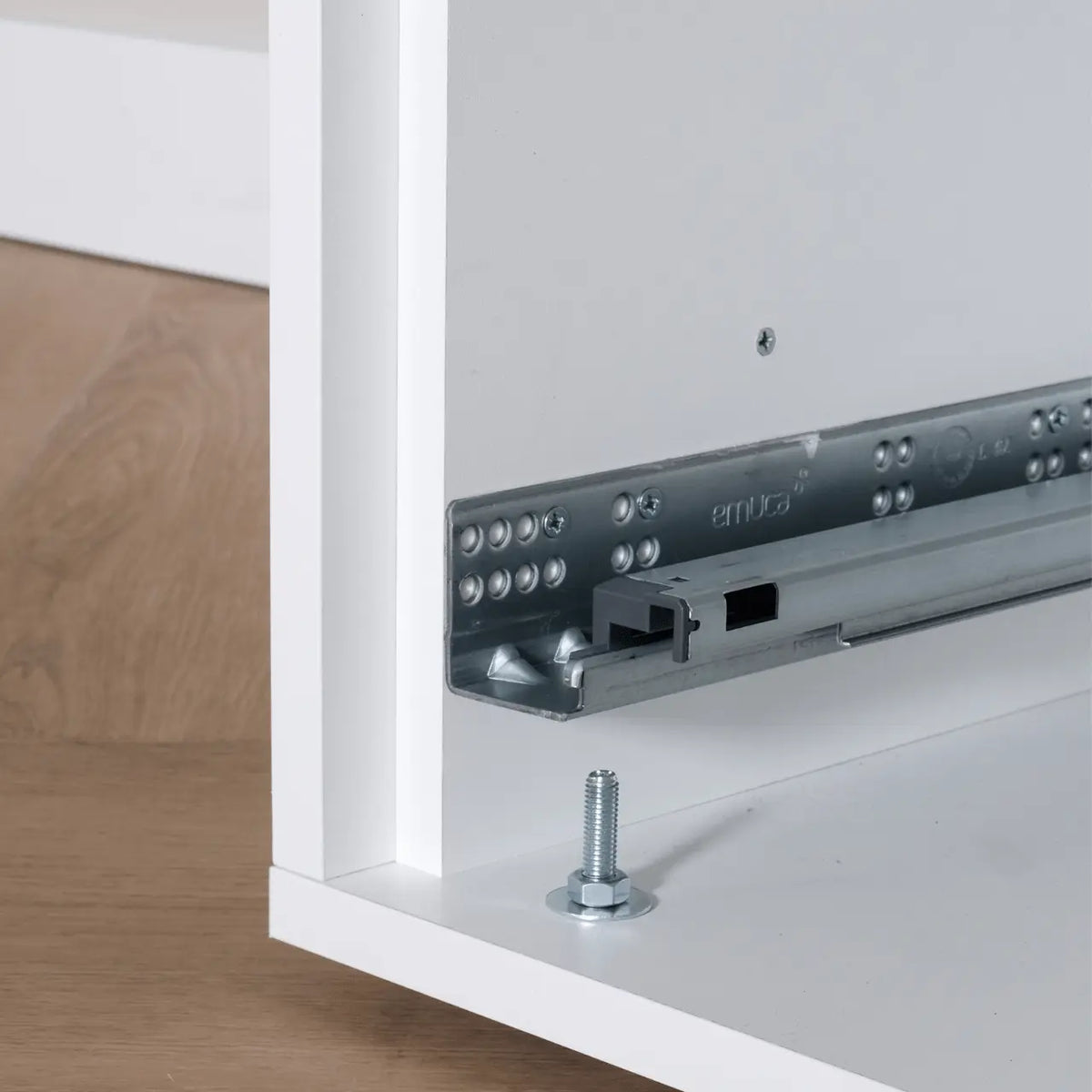 Metal drawer slide rail and white wooden cabinet corner with mounting bolt on wood floor background.
